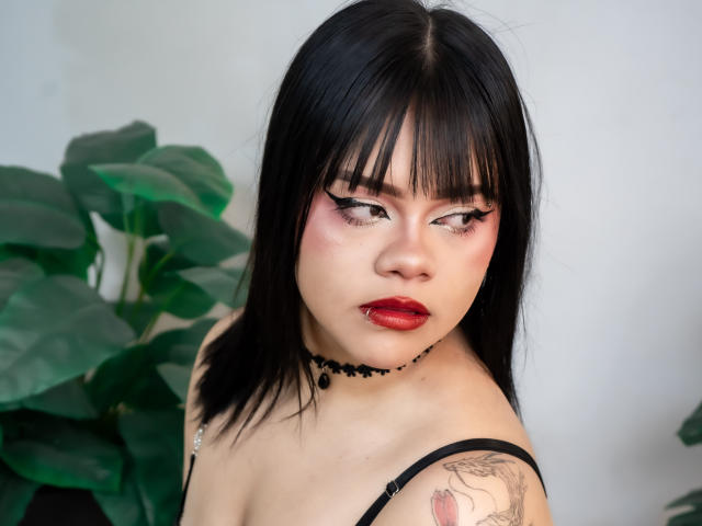 AnneStonee has black hair, red lipstick, and eyeliner, wearing a black choker and strap, with a tattoo on her upper arm, looking sideways with a serious expression.