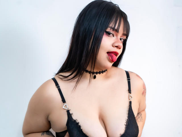 AnneStonee with black hair, red lipstick, and black choker, sticking out tongue, wearing black lace bra, showing large tits, tattoos on arms, white background.