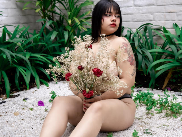 AnneStonee, topless with small tits, black panties, holding flowers, sitting on white gravel, green plants, red and white flowers, tattoo on right arm, short black hair, looking at camera.