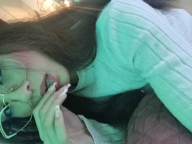 MadisonSmitt with long brown hair, green ribbed sweater, and glasses, biting her pink manicured finger, lying on a dark surface with a pink quilt.