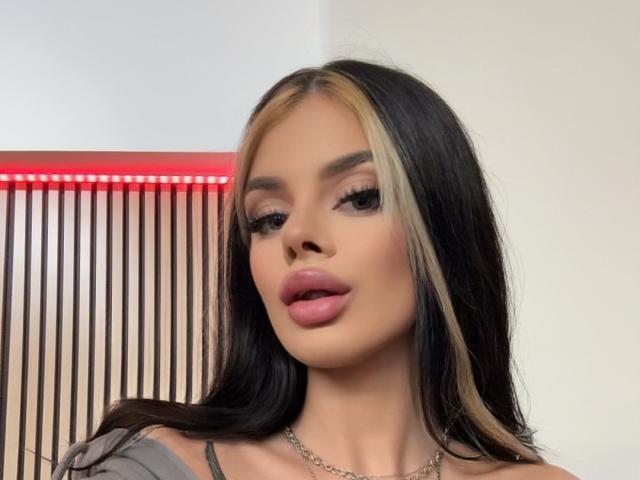 IvyRaven has long black hair with blonde highlights, full pink lips, and a silver necklace. She's looking at the camera with a slight pout. Red LED strip behind.
