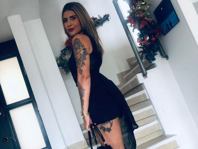 SofiFoxx with tattoos on her arm and thigh, wearing a black dress, standing on stairs, holding black heels, looking back with red lipstick, and a smile.
