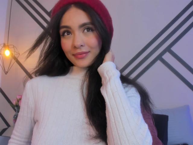 MadisonSmitt with long black hair, wearing a red hat and white ribbed sweater, smiling slightly, holding her hair up. Soft light, warm tones, and a subtle pink blush.