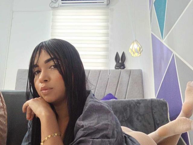 AliceSmity, a young woman with long black hair and brown skin, lies on a grey couch with her head resting on her hands. She wears a loose black shirt and beige fishnet socks. Her right arm has a gold bracelet. She has a neutral expression, looking slightly to the right. The background includes a white window with blinds, a hanging geometric yellow light, and a purple geometric wall. A black bunny head decorates the wall behind her. Her legs are bent at the knees, feet up in the air.