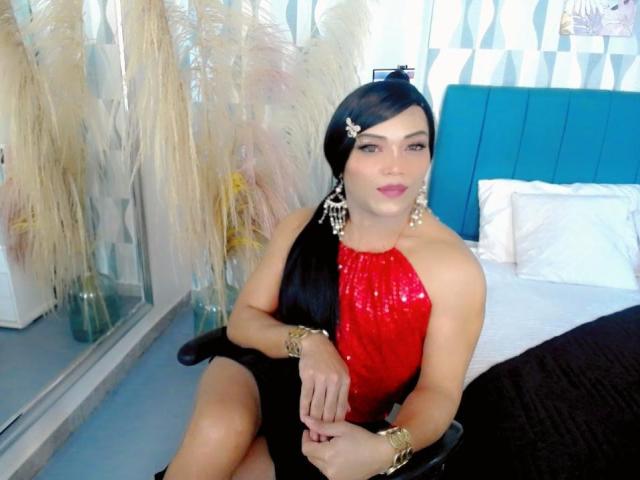 CharlotteSweetX with long black hair, red halter dress, gold earrings, black skirt, and gold bracelet, sitting on bed, looking at camera, smiling, puffy white pillows in background.