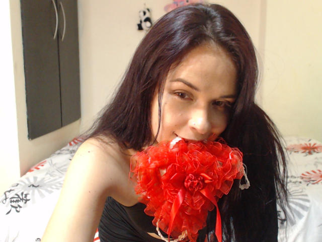 Wendy Schulz, avec ses cheveux brun foncé, tenant une rose rouge et duveteuse dans sa bouche, souriant, portant un haut noir, laissant apparaître ses épaules nues et une légère peau bronzée..