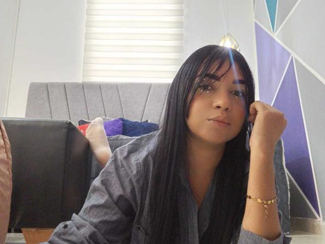 Alice Smity, met lang zwart haar, draagt een grijze blouse, met haar hoofd rustend op haar hand, kijkt naar de camera, heeft een kleine gouden armband om haar pols, een subtiele glimlach, zacht licht..