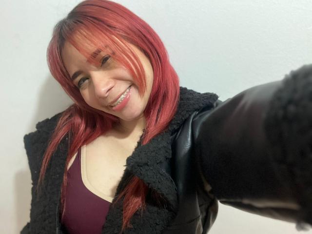 MelannyTaylorr with red hair, smiling, wearing a black fuzzy jacket and maroon bra, taking a selfie with her right arm, showing her tits slightly.