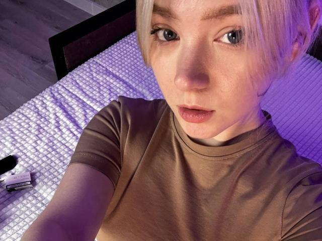 EnchantingEyes with short blonde hair, light skin, and blue eyes, wearing a brown turtleneck, has a slightly open mouth, and is lying on a white textured bed.