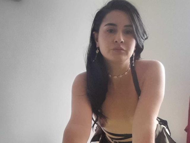 FrancheskaWaalsh with black hair, wearing a halter top, showing her tits, looking down, arms slightly forward, wearing a necklace, in a softly lit room.