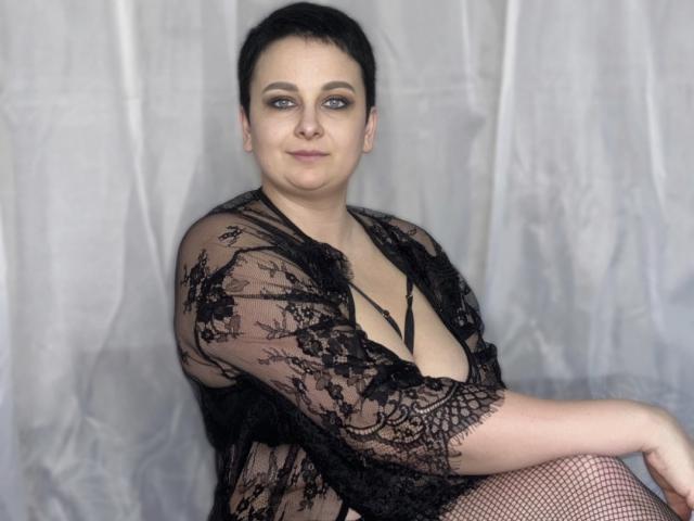 CelinaGinny, black lace robe, deep cleavage, fishnet stockings, short black hair, light skin, slight smile, sitting, blue eyes, looking at camera, soft light, subtle makeup.
