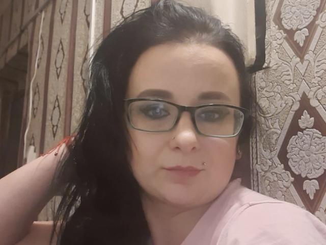CelinaGinny with black hair, glasses, nose piercing, and pink shirt, looking at camera with a slight smile, in a room with patterned wallpaper. Her ass is not visible.