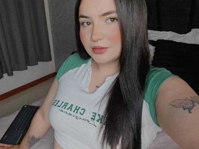 ValeriJoness has long black hair, green eyes, and a butterfly tattoo on her right arm. She's wearing a green and white raglan shirt, sitting with a remote in hand.