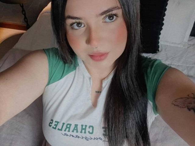 ValeriJoness with long black hair, green eyes, and a nose ring, wearing a green and white raglan shirt, showing her small tits and a tattoo on her right arm.