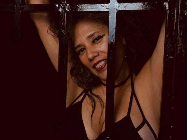 CuteMatureSassy, with curly dark hair and red lipstick, smiling through black metal bars, wearing a black strappy top, exposing her tits, arms raised, looking sexy and confident.