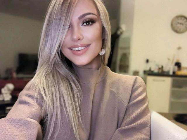 VickyFox, aantrekkelijk met lang blond haar, lachend, met een coltrui, haar neusring zichtbaar en roze lippen, in een gezellige, overdekte omgeving, met roze oorbellen..