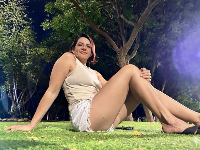 MissSofy with red hair, sleeveless beige top, and white frayed shorts, sitting on grass, legs crossed, smiling, arms behind, bare feet, night, trees in background.