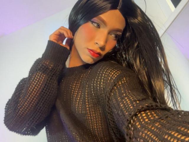 DominiqueR with long black hair, red lipstick, and a black mesh sweater, showing off her tits and looking at the camera with a slight smirk.