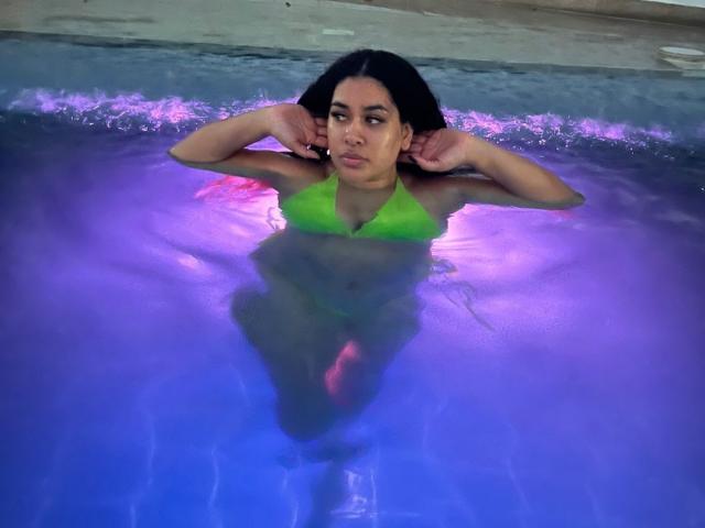 LianaAndAsaff with dark hair, green bikini top, and wet skin, floating in a purple-tinted pool, arms behind head, looking off to the side with a serious expression.