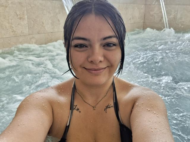 KatherinLatin with wet black hair, small tits, tattoos on chest, smiling in a hot tub, water droplets on skin, wearing a black halter top, taking a selfie.