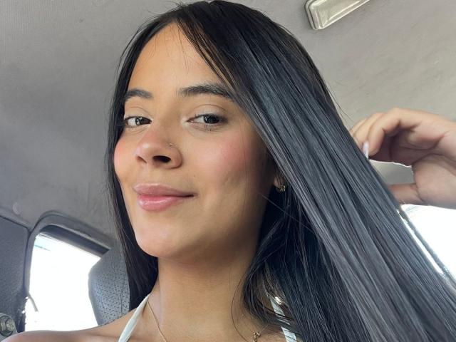 TamaraPeach has long, straight black hair, a small nose piercing, and is smiling slightly with pink lips. She's holding her hair with one hand, showing off her cute face.