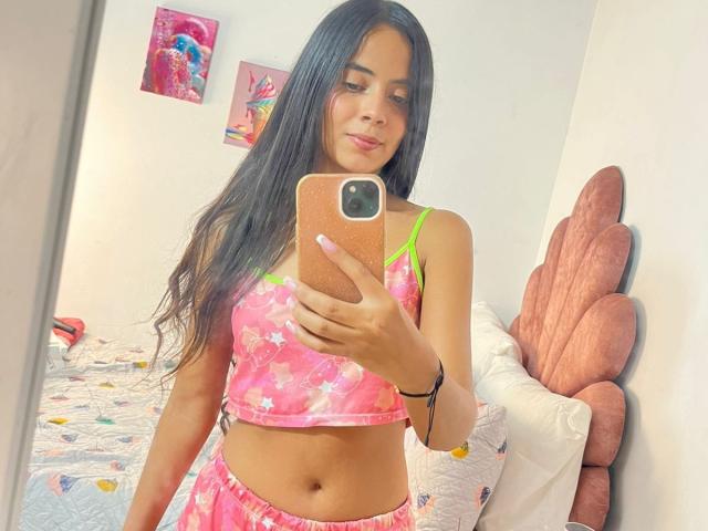 TamaraPeach with long black hair, wearing a pink floral crop top and matching skirt, holding a pink phone, mirror selfie, showing her small tits and flat ass, in a bright room.