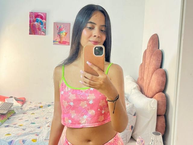 TamaraPeach, a young woman with long black hair, takes a mirror selfie. She wears a pink floral crop top with green straps and matching pink shorts. She holds an orange phone with a black case. Her pink nails are manicured. She has a slight smile. The background includes a pink, scallop-patterned headboard, a white pillow, and colorful wall art. The bed has a patterned quilt. The image is bright with natural light.
