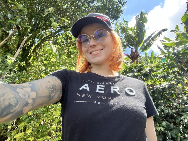SarahRousen with red hair, wearing a black cap, reflective sunglasses, and a black t-shirt, smiling, showing her tattooed arm, and her small, perky tits.