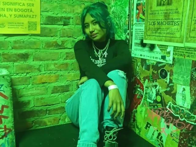 NovaAllen, con cabello azul, camisa negra y jeans azules, sentada con las piernas cruzadas, usando un collar y una pulsera, foto con un tono verdoso, mirando ligeramente hacia un lado..