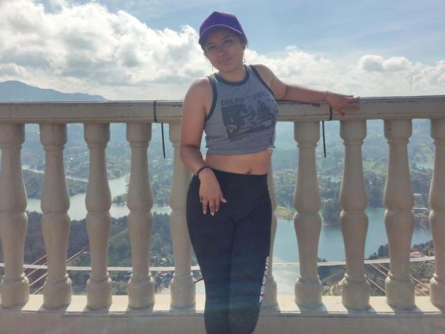NovaAllen stands with a hand on a balustrade, wearing a gray tank top showing her tits and black pants, purple cap, and bracelet, with a mountain view behind.