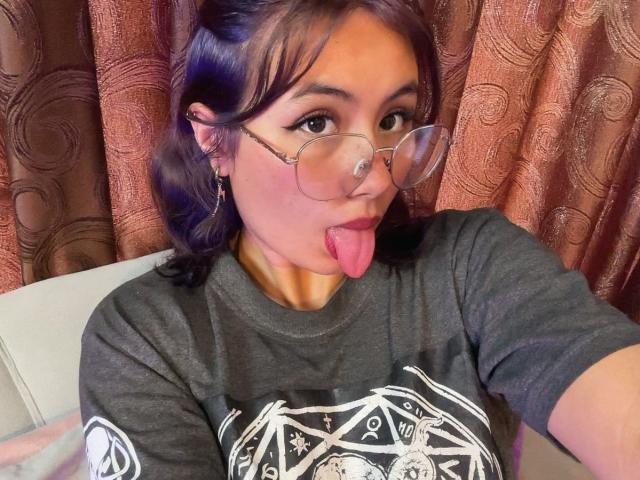 NovaAllen, com cabelo roxo, óculos e piercing no nariz, mostrando a língua, vestindo uma camisa cinza escura com um desenho branco, tirando uma selfie mostrando seu rosto..