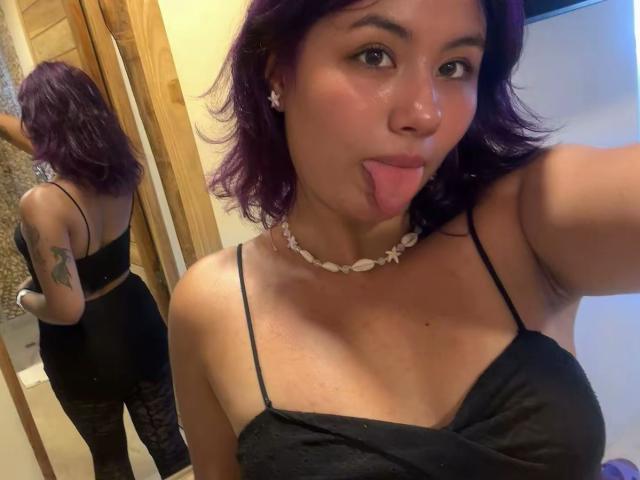 NovaAllen with purple hair, black spaghetti top, and choker, sticking her tongue out, showing her small tits and black lace-pantied ass in a mirror selfie.