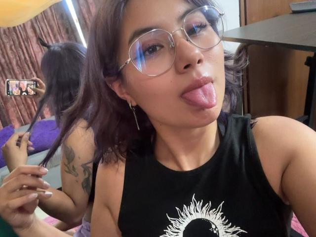 NovaAllen with dark purple hair, glasses, and black sleeveless top, sticking out her tongue, showing a tattoo on her right arm, and holding a phone in a mirror selfie.