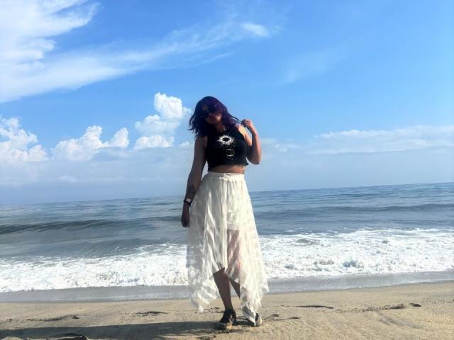 NovaAllen tem cabelos longos e escuros, usa um boné preto com caveira e uma saia branca, transparente e fluida, estando em pé em uma praia arenosa com ondas ao fundo..