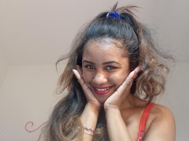 A close-up photograph of LayahCoquine. She has long, wavy brown hair with a blue hair tie, smiling with hands on her cheeks. She has a medium-brown skin tone, red lipstick, and is wearing a red strap top. Her fingers have silver rings. Her eyes are brown, and her hair is slightly messy. The image has a soft, natural light. Her expression is playful and friendly.