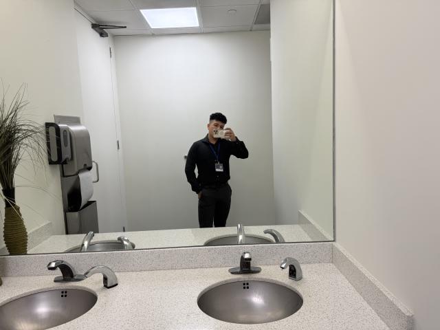 LeoBlaze, wearing a black cap and shirt with hands in pants, stands in front of a mirror, taking a selfie with a phone in a clean bathroom.