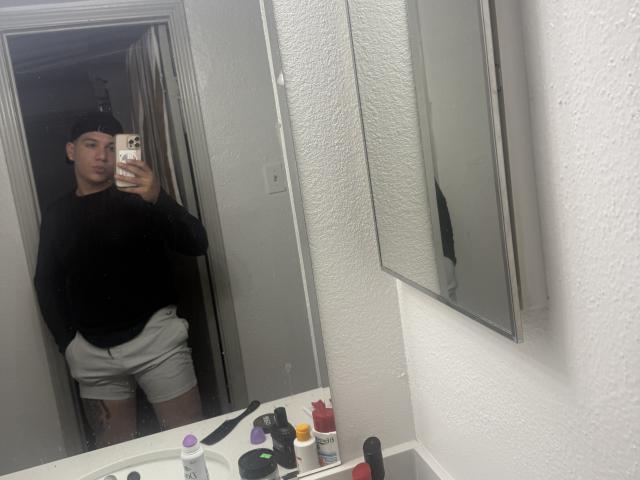 LeoBlaze stands in a bathroom, wearing a black sweater and gray shorts, taking a mirror selfie. His phone is in his right hand, with a white wall behind.