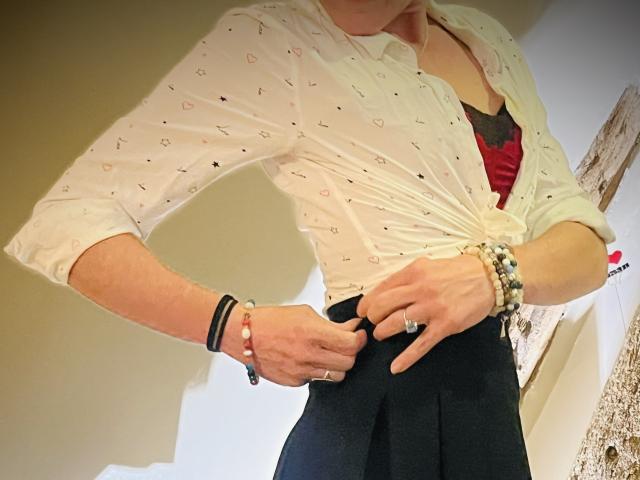 TifolyLaurent in a white, patterned, button-up shirt tied at the waist, black skirt, showing a red bra, wearing multiple bracelets and rings, adjusting her skirt.