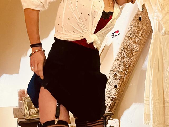 TifolyLaurent's white, button-up shirt tied at the front, black skirt, red bra, garter, and black stockings with a visible thong. Her hand's on her skirt's side.