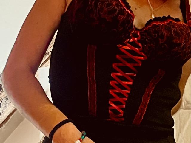TifolyLaurent in black lace and red corset with red ribbon, showing tits, wearing a black bracelet and green bead bracelet, hand on hip.