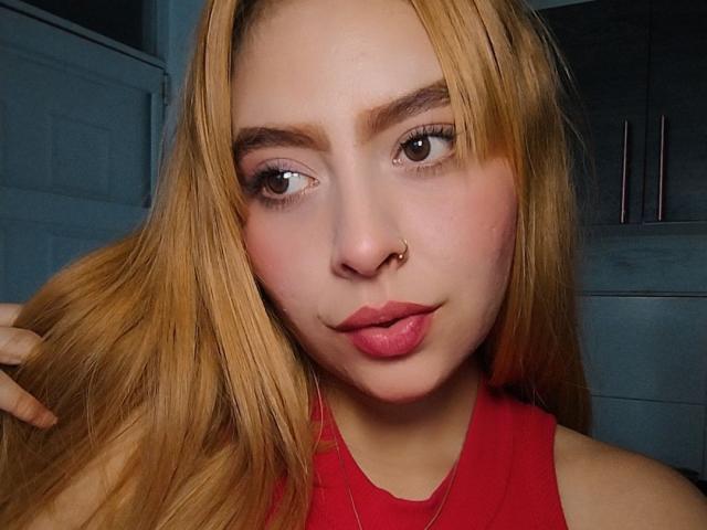LolaEvans with blonde hair, nose ring, and pink lips, pouting slightly, wearing a red top, looking at the camera, holding her hair on the left.