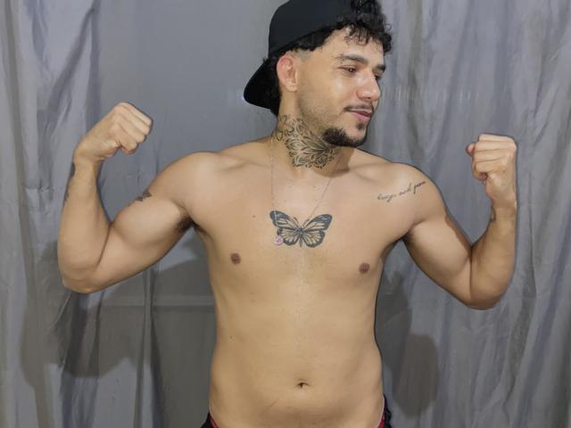 Felai, un homme latino musclé avec des cheveux noirs bouclés, portant une casquette noire, contracte ses bras, montrant un tatouage de papillon sur sa poitrine et des tatouages sur son cou et son épaule..
