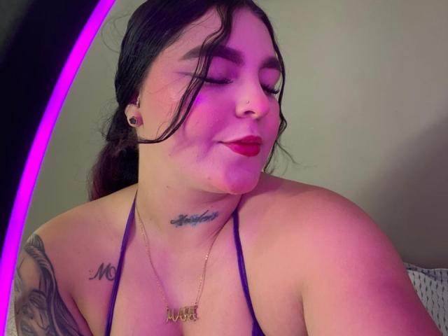MelodyBlossom hat einen rosafarbenen Hautton, dunkles Haar und lächelt mit geschlossenen Augen. Sie trägt ein violettes Halstopp, das Tattoos an ihrem Schulter- und Nackenbereich zeigt, und hat einen Nasenring..