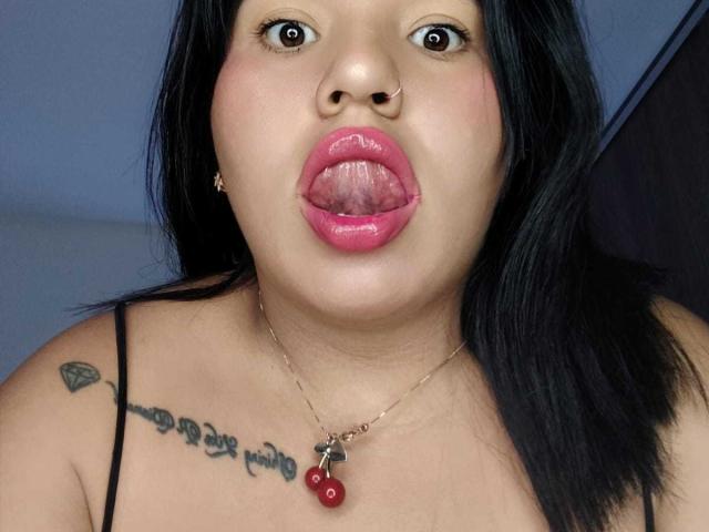 CristinaMendez with black hair, nose ring, and pink lipstick, tongue out, looking up, chest tattoo, necklace with red cherries, showing her tits.