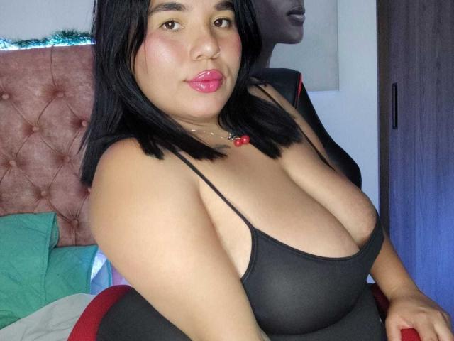 CristinaMendez has black hair, pink lipstick, and large tits in a black, sheer top. She's sitting, showing her curvy, sexy body, with a red, button-up chair.