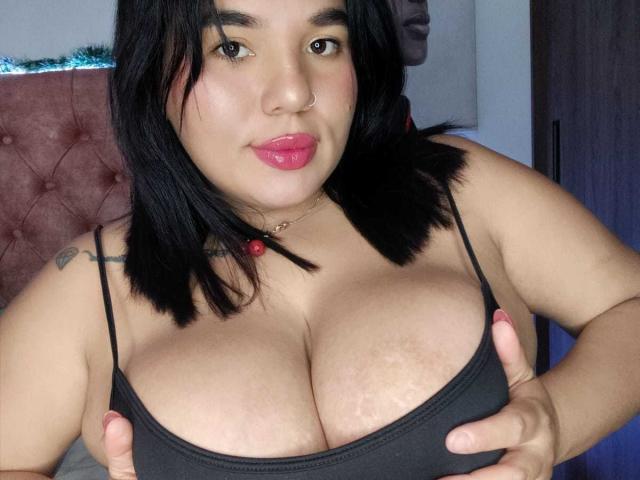 CristinaMendez has big, round tits, wearing a black top, with hands on her chest, black shoulder-length hair, pink lips, nose ring, and tattoo on her right shoulder.