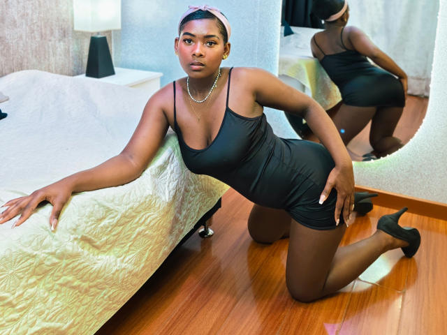KarinaFlowers, dark-skinned, in black dress, kneeling on wooden floor, showing her ass, small boobs, in mirror's reflection, with pink headband, pearl necklace, black heels, and white bed.