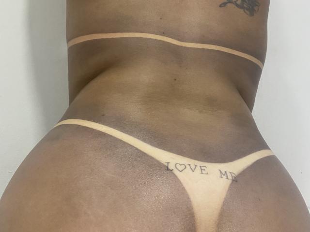 AylaSafadinha's tan thong covers her ass, showing a tattoo that reads "LOVE ME" in black letters. She has dark brown skin with a tattoo on her lower back.