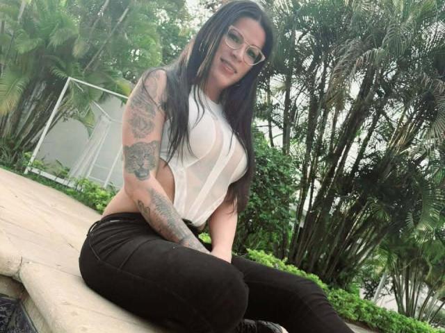 SexyCaandydoll with long black hair, glasses, white semi-sheer top showing tits, black pants, sitting on stone ledge, tattoos on left arm, smiling, lush green outdoor background.