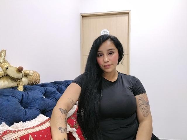 Shariit, met lang zwart haar, gekleed in een zwarte blouse, zittend op een blauwe gestoffeerde bank, met tatoeages op haar armen zichtbaar, kijkt naar de camera met een lichte glimlach..
